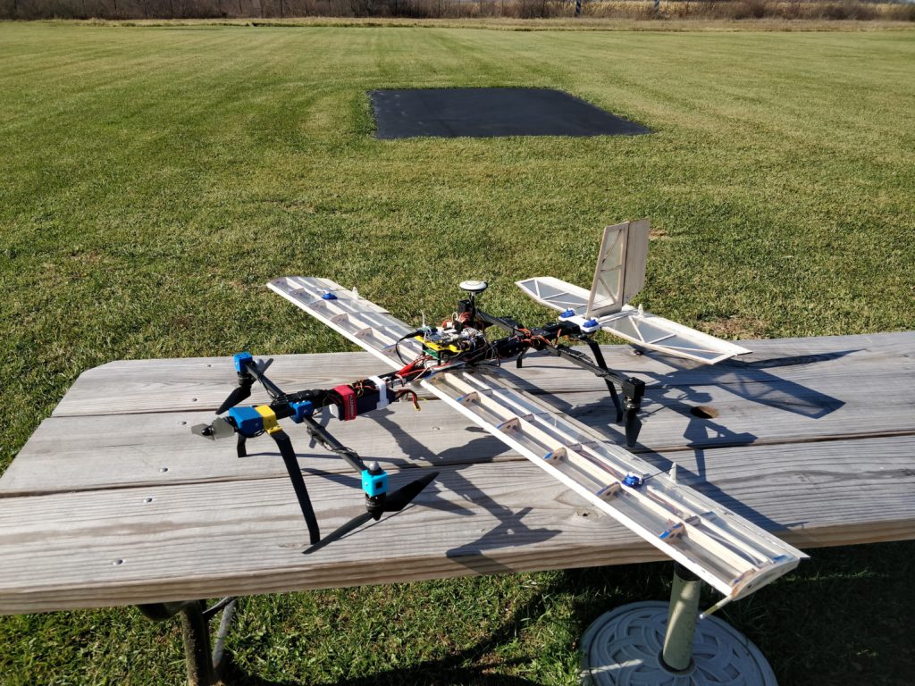 A hybrid quadplane with fixed wings and vertical-lift propellers on a table at a flying field.